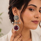 A close-up of a woman wearing a large silver-toned statement earring with a round purple gemstone and pearl-like accents, against a soft beige background.