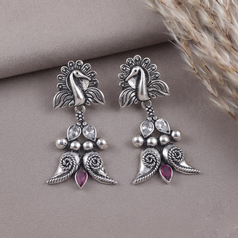 A pair of silver-toned dangle earrings with a peacock motif, featuring clear teardrop crystals and small pink teardrop gemstones, displayed on a textured taupe surface with dried grass in the corner.