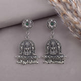 A pair of silver-toned Indian jhumka earrings with detailed Hamsa motifs, small green stone accents in the studs, and dangling bead chains, displayed on a neutral gray background with dried grass.