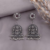 A pair of ornate silver-toned Indian jhumka earrings with detailed deity carvings and small red gemstone accents, displayed on a neutral gray background with dried grass.