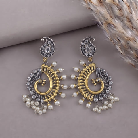 A pair of ornate Indian-style dangle earrings with gold and silver-tone metalwork, featuring paisley tops, fan-shaped details, and clusters of white faux pearls, displayed on a neutral gray surface with dried grass in the corner.