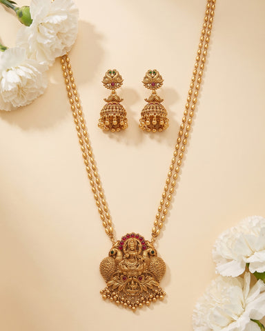 Gold-toned traditional jewelry set featuring a necklace with an ornate pendant and matching drop earrings, arranged on a warm beige background with white flowers in the corners.