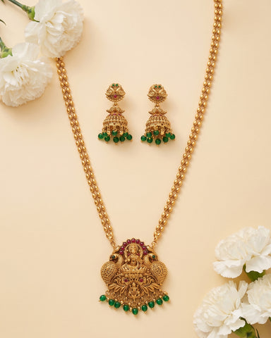Gold-toned traditional necklace and matching earrings with green stone accents and a central deity pendant, arranged on a warm beige background with white flowers in the corners.