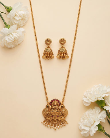 Gold-toned traditional jewelry set featuring a necklace with a detailed peacock and deity pendant, matching drop earrings, and white flowers arranged on a light beige background.