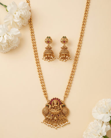 Gold-toned traditional necklace and matching dangle earrings with pearl-like beads and small red and green stone accents, arranged on a warm beige background with white flowers in the corners.