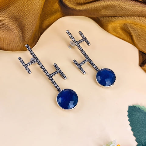 A pair of silver-toned geometric earrings with round deep blue gemstones, displayed on a beige surface with draped gold fabric in the background.