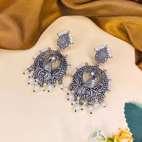 A pair of ornate silver-toned statement earrings with carved peacock motifs and dangling white pearl beads, displayed on a beige surface with draped gold fabric in the background.