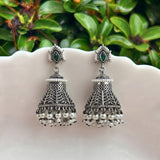 A pair of traditional Indian silver-toned jhumka earrings with green stone accents and dangling pearl-like beads, displayed on a white scalloped dish with green foliage in the background.