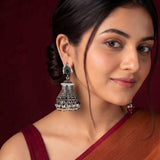 A woman wearing a traditional Indian silver jhumka earring with green and white stones, shown in a close-up portrait against a dark red background.