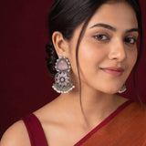 A close-up of a woman wearing ornate silver-toned jhumka earrings with pink stones and white pearl accents, against a dark burgundy background.