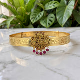 An ornate gold-toned traditional Indian bangle bracelet featuring a central Ganesha medallion and dangling red gemstone beads, displayed on a white marble surface with green plants in the background.
