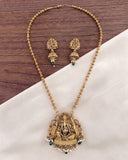 Gold-toned Ganesha necklace and matching earrings displayed on a white fabric surface with a dark wood background. The jewelry features an ornate Ganesha pendant and matching drop earrings with small green and white stone accents.