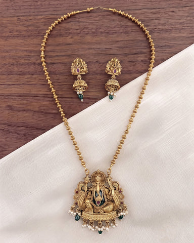 Gold-toned Ganesha necklace and matching earrings displayed on a white fabric surface with a dark wood background. The jewelry features an ornate Ganesha pendant and matching drop earrings with small green and white stone accents.