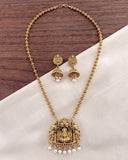 Gold-toned traditional necklace and matching earrings with ornate pendant details, pearl drops, and dark stone accents, arranged on a white fabric surface over a dark wood background.