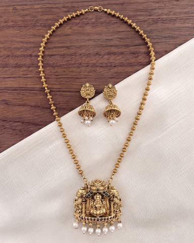 Gold-toned traditional necklace and matching earrings with ornate pendant details, pearl drops, and dark stone accents, arranged on a white fabric surface over a dark wood background.