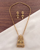 Gold-toned traditional Indian jewelry set featuring a detailed temple-style pendant necklace and matching earrings, displayed on a white fabric surface over a dark wood background.
