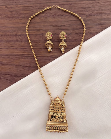 Gold-toned traditional Indian jewelry set featuring a detailed temple-style pendant necklace and matching earrings, displayed on a white fabric surface over a dark wood background.