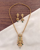 Gold-toned jewelry set featuring a necklace and matching earrings with a detailed pendant depicting a deity, accented with green stones and small white bead details, displayed on a white fabric and dark wood background.