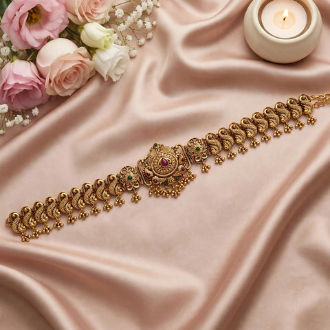 An ornate gold-toned traditional Indian necklace with intricate paisley motifs and small red and green stone accents, displayed on draped blush pink satin fabric with pink and white roses and a lit candle in the background.
