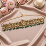 An ornate gold-toned traditional necklace with intricate filigree, red and green stone accents, and a row of dangling green beads, displayed on a draped pink satin fabric with roses and a candle.