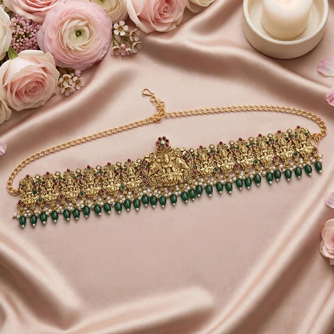 An ornate gold-toned traditional necklace with intricate filigree, red and green stone accents, and a row of dangling green beads, displayed on a draped pink satin fabric with roses and a candle.
