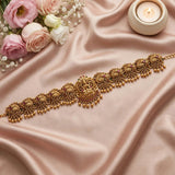 An ornate gold-toned traditional Indian choker necklace with intricate filigree, bead drops, and small pink stone accents, displayed on draped blush satin fabric with roses and a lit candle in the background.