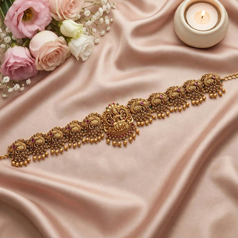 An ornate gold-toned traditional Indian choker necklace with intricate filigree, bead drops, and small pink stone accents, displayed on draped blush satin fabric with roses and a lit candle in the background.