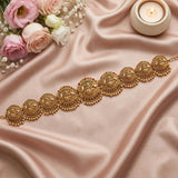 An ornate gold-toned traditional waist chain or belt with intricate filigree and bead details, arranged diagonally on draped blush pink satin fabric, with pink and white roses in the top left and a lit candle in the top right.
