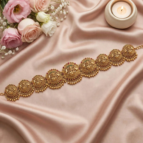 An ornate gold-toned traditional waist chain or belt with intricate filigree and bead details, arranged diagonally on draped blush pink satin fabric, with pink and white roses in the top left and a lit candle in the top right.