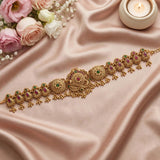 An ornate gold-toned traditional Indian choker necklace with intricate filigree work, red and green stone accents, and dangling bead details, displayed on draped blush pink satin fabric with pink and white roses and a lit candle in the corners.
