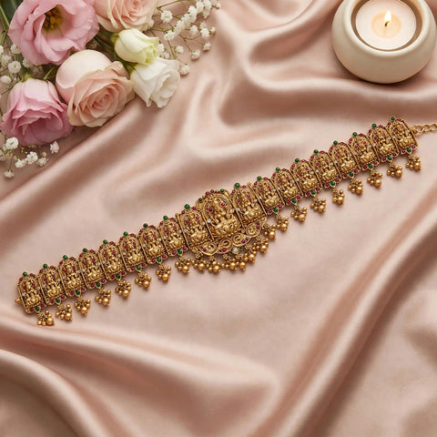 An ornate gold-toned traditional Indian choker necklace with detailed filigree and small green stone accents, displayed on draped blush pink satin fabric with pink and white roses and a lit candle in the background.