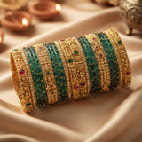 A stack of traditional Indian bangles, featuring intricate gold-plated filigree and green stone-encrusted bands, arranged on draped beige silk fabric with warm diya lights blurred in the background.