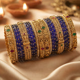 A stack of ornate Indian bangles, including gold-plated filigree bands with red and green stone accents and deep blue beaded bangles, arranged on draped satin fabric with blurred diya candles in the background.