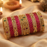 A stack of traditional Indian bangles, including bright pink beaded bracelets and ornate gold-plated rings with green and red stone accents, displayed on draped beige fabric with blurred diya candles in the background.