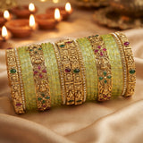 A set of traditional Indian bangles, including light green stone-wrapped bands and ornate gold-plated filigree bangles with red and green stone accents, displayed on draped satin fabric with blurred diya candles in the background.