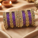 A stack of traditional Indian bangles, including purple beaded bangles and ornate gold-plated bangles with red and green stone accents, displayed on draped satin fabric with blurred diya candles in the background.