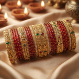 A stack of ornate Indian bangles (kara bracelets) featuring red glass beads, gold-plated metal bands, and small green and red stone accents, displayed on draped beige fabric with blurred diya candles in the background.