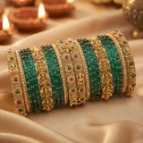 A stack of traditional Indian bangles, featuring green stone-wrapped bands alongside ornate gold-plated filigree bangles with red and green stone accents, displayed on draped beige fabric with blurred diya candles in the background.