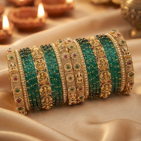 A stack of traditional Indian bangles, featuring green stone-wrapped bands alongside ornate gold-plated filigree bangles with red and green stone accents, displayed on draped beige fabric with blurred diya candles in the background.