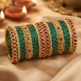 A stack of traditional Indian bangles (boca bracelets) featuring emerald green beaded stripes, gold-plated borders, and red gemstone accents, displayed on a draped beige satin fabric with blurred diya candles in the background.