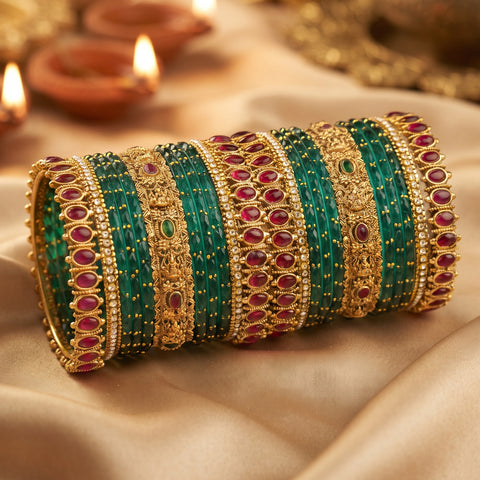 A stack of traditional Indian bangles (boca bracelets) featuring emerald green beaded stripes, gold-plated borders, and red gemstone accents, displayed on a draped beige satin fabric with blurred diya candles in the background.