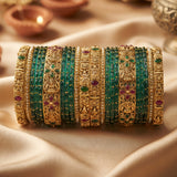 A set of traditional Indian bangles (kara) displayed in a row. The set features gold-plated bangles with intricate filigree patterns, some adorned with small green and red stones, alongside multiple thin green beaded bangles.