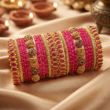 A stack of traditional Indian bangles, including bright pink stone-wrapped bracelets and ornate gold-plated bangles with green and white stone accents, displayed on draped beige satin fabric with blurred diya lights in the background.