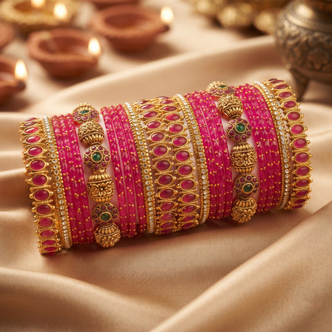 A stack of traditional Indian bangles, including bright pink stone-wrapped bracelets and ornate gold-plated bangles with green and white stone accents, displayed on draped beige satin fabric with blurred diya lights in the background.