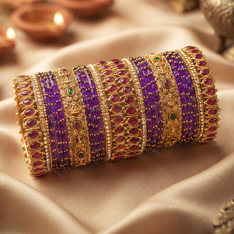 A stack of traditional Indian bangles, featuring purple beaded bracelets alongside ornate gold-plated bangles embellished with red stones and small green accents, displayed on draped beige satin fabric with warm background bokeh.