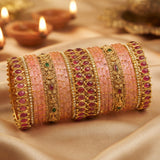A stack of traditional Indian bangles, including pink beaded bracelets and gold-plated rings with red and green stone accents, displayed on a draped beige satin fabric with blurred diya candles in the background.