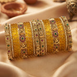 A stack of traditional Indian bangles, including solid yellow glass bangles and gold-plated bangles decorated with red, green, and blue stones, displayed on draped beige satin fabric.