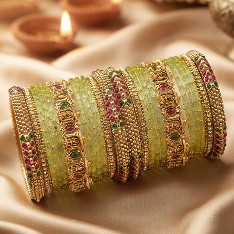 A stack of ornate Indian bangles (brocades) made of light green stone inlays, gold-plated metal, and embellished with red and green gemstones, displayed on draped beige satin fabric with blurred diya candles in the background.