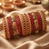 A set of traditional Indian bangles (brocaches) made of gold-plated metal, decorated with red stones, small gold beads, and a few green stone accents, arranged on a draped beige satin fabric with warm background lights.