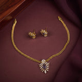 Gold jewelry set featuring a necklace and matching earrings, displayed on a dark burgundy fabric background with a wooden surface in the corner. The necklace has a braided gold chain and a central leaf-shaped pendant with a dark teardrop stone and small clear stones.
